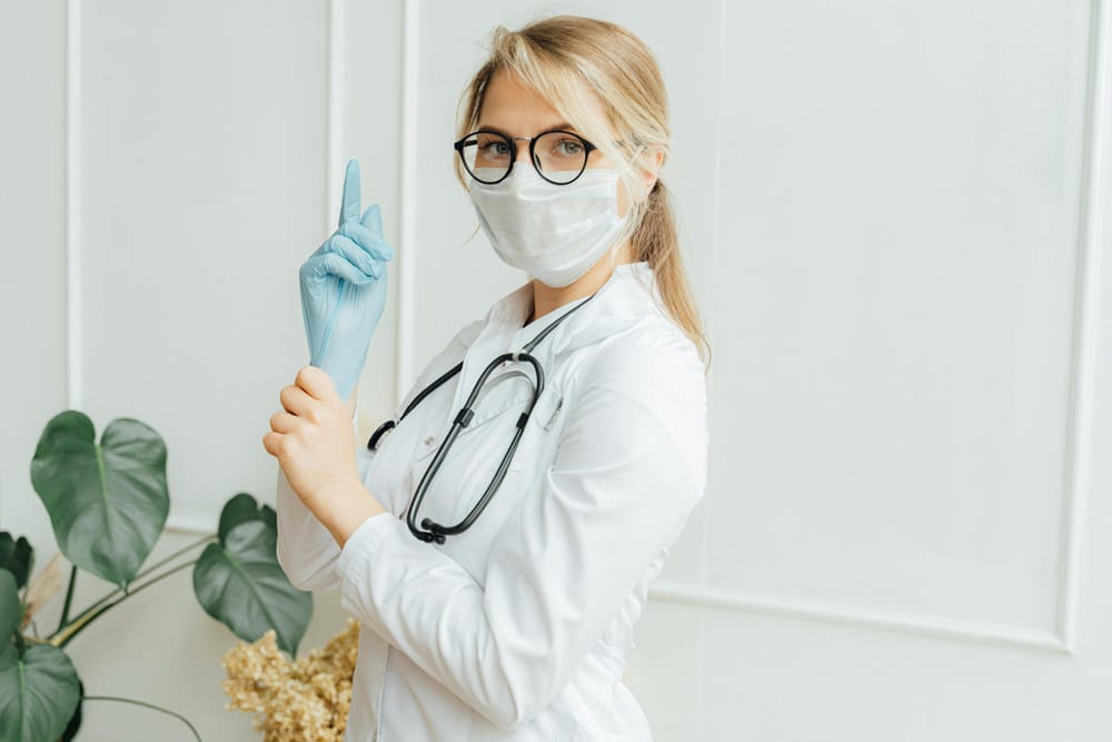 Blonde doctor, in white lab coat, putting on blue latex gloves.