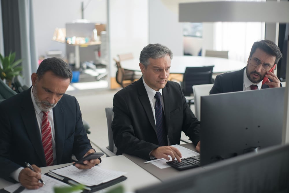 Three men sitting at a desk in front of a computer with paperwork concerning fema relief funding in front of them.