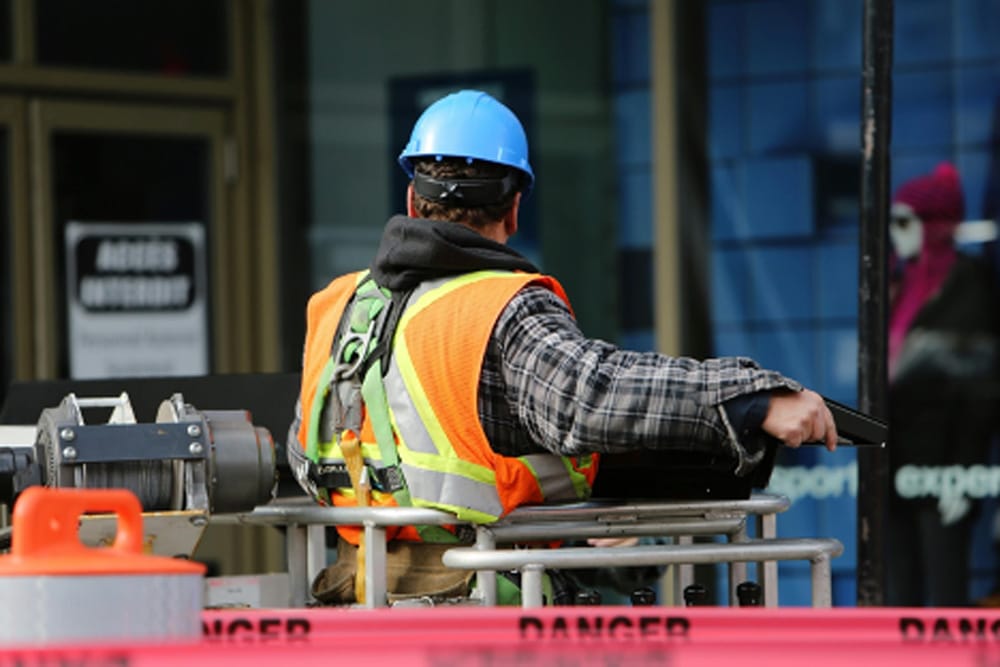 Man in an orange safety vest and blue hardhat standing on equipment.