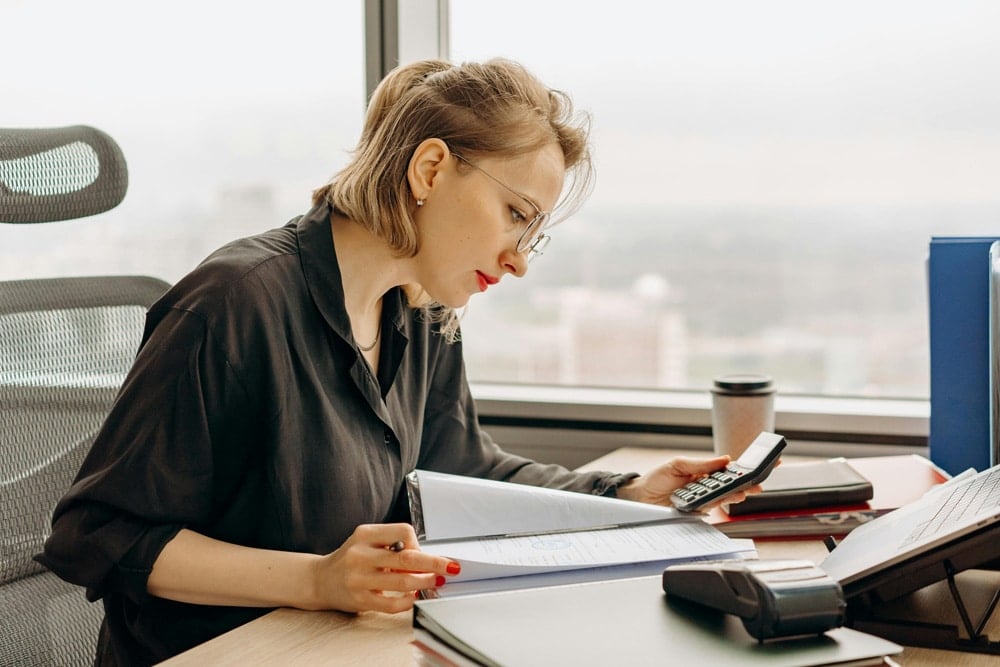 A focused woman reviews documents with a pen and calculator at a desk in a bright office, working on disaster financing tasks. A cityscape is visible outside.