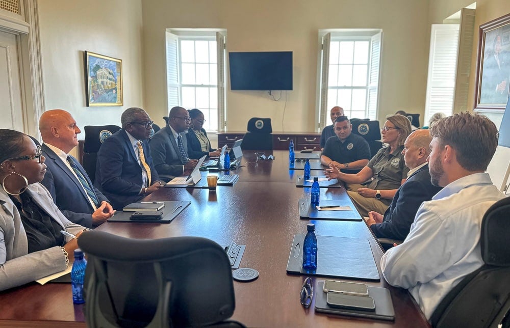 A diverse group in a formal meeting room discusses disaster financing around a large table with laptops, notebooks, and water bottles.