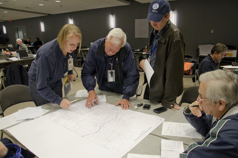 People standing around paperwork on a wooden desk discussing a debris management plan after a disaster.